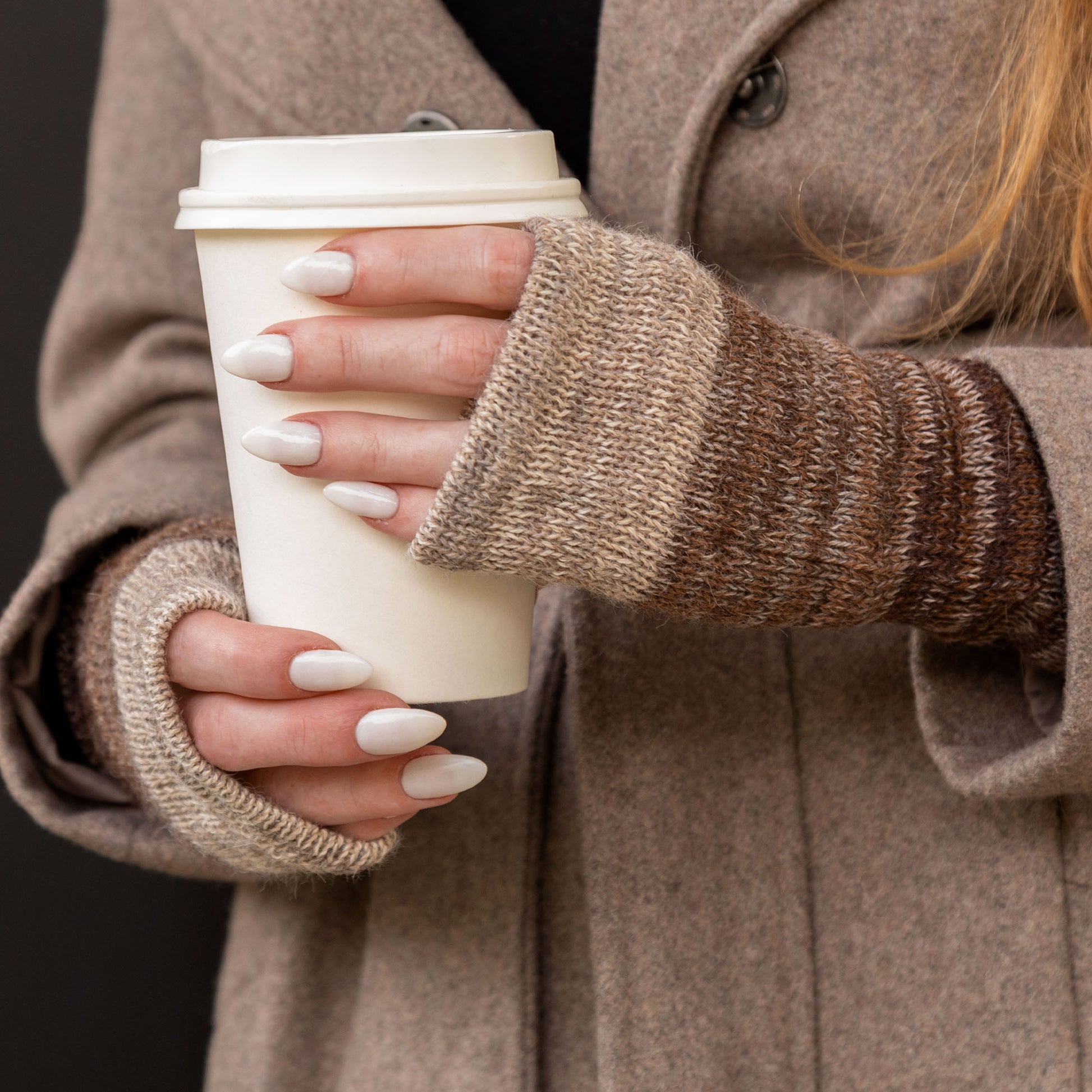 Person holding a white paper coffee cup while wearing brown fingerless mittens with a brown coat