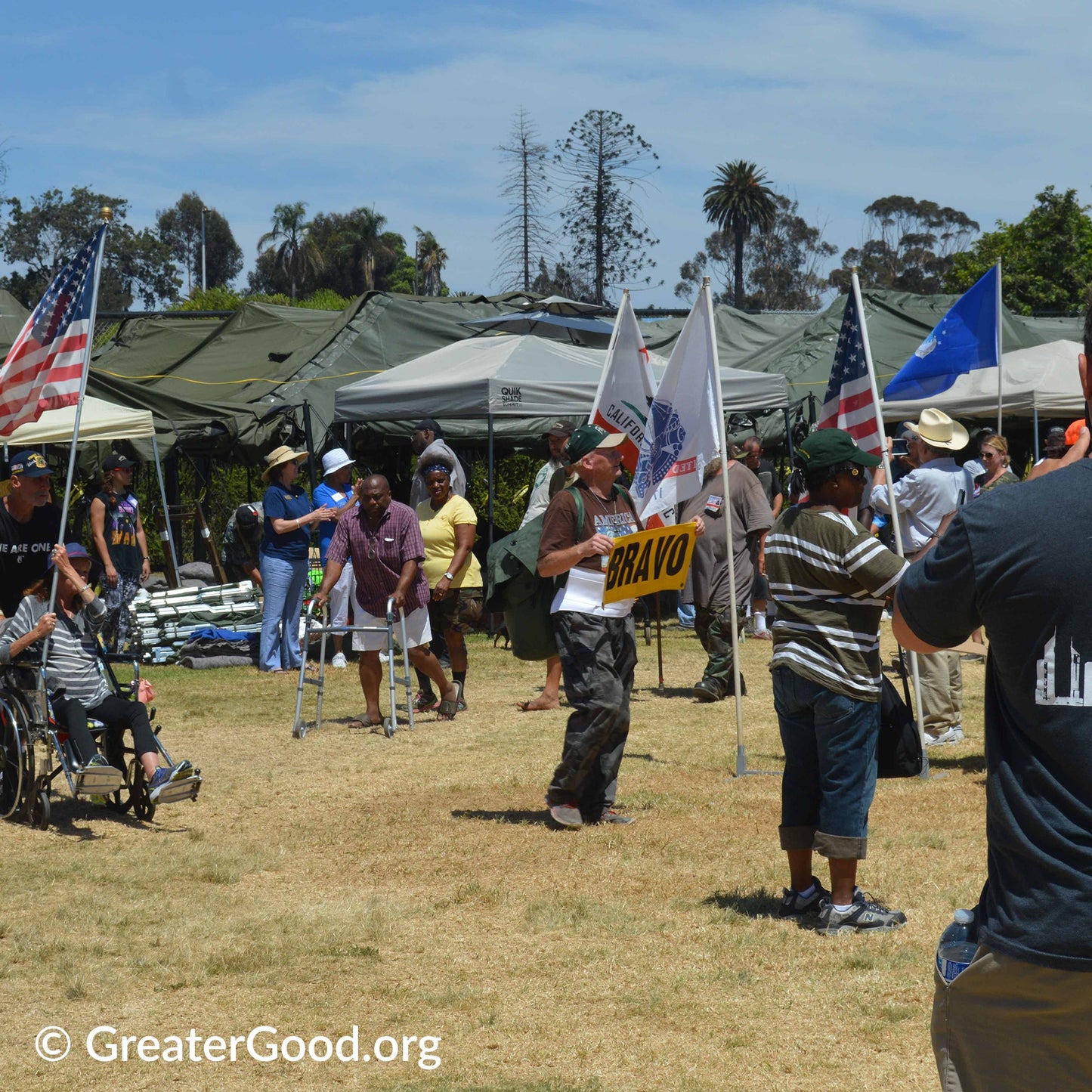 Outdoor event with people and tents, including a person holding a 'Bravo' sign.