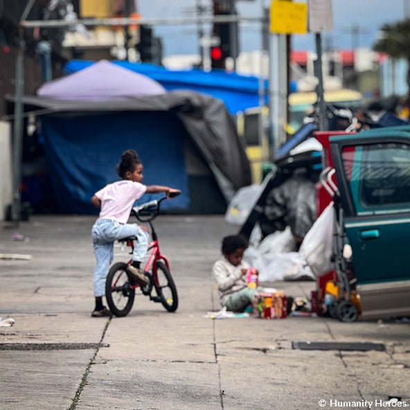 Child on a bike near a homeless encampment with tents and people sitting on the ground.