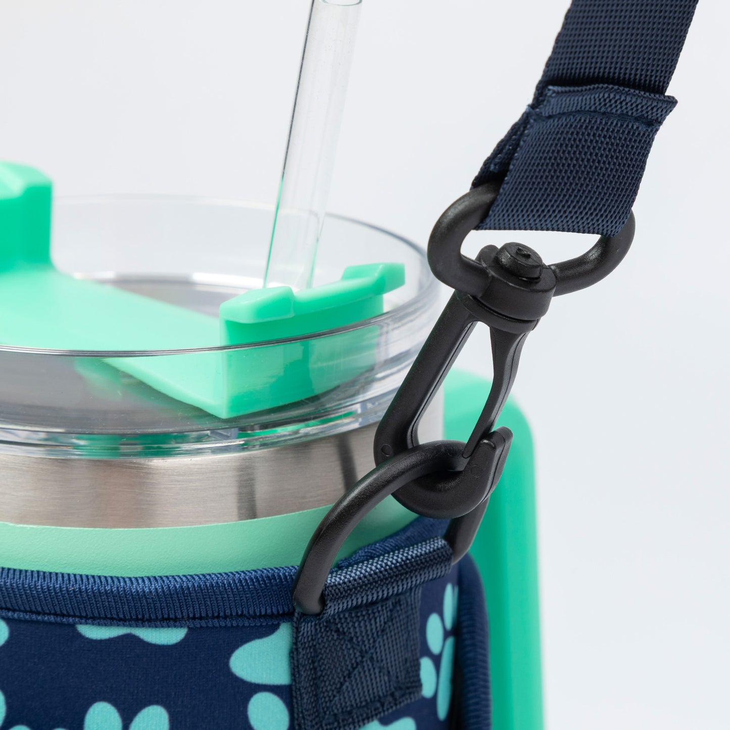 Close-up of a insulated tumbler with a clear straw, mint green lid and handle, and a navy blue paw-print patterned carrying sleeve with a black carabiner strap.