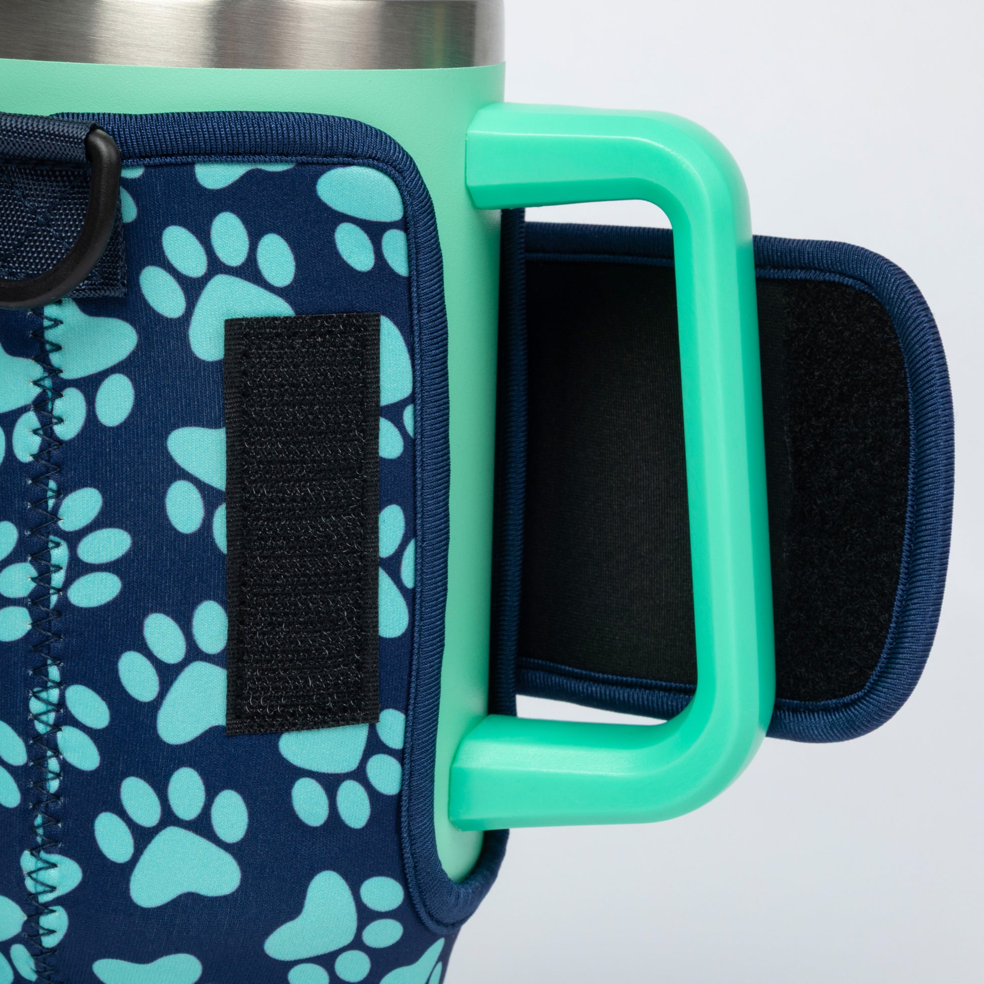Close-up of a handled insulated mug with a mint green handle and rim, a navy sleeve printed with light blue paw prints, and a black hook-and-loop closure panel.