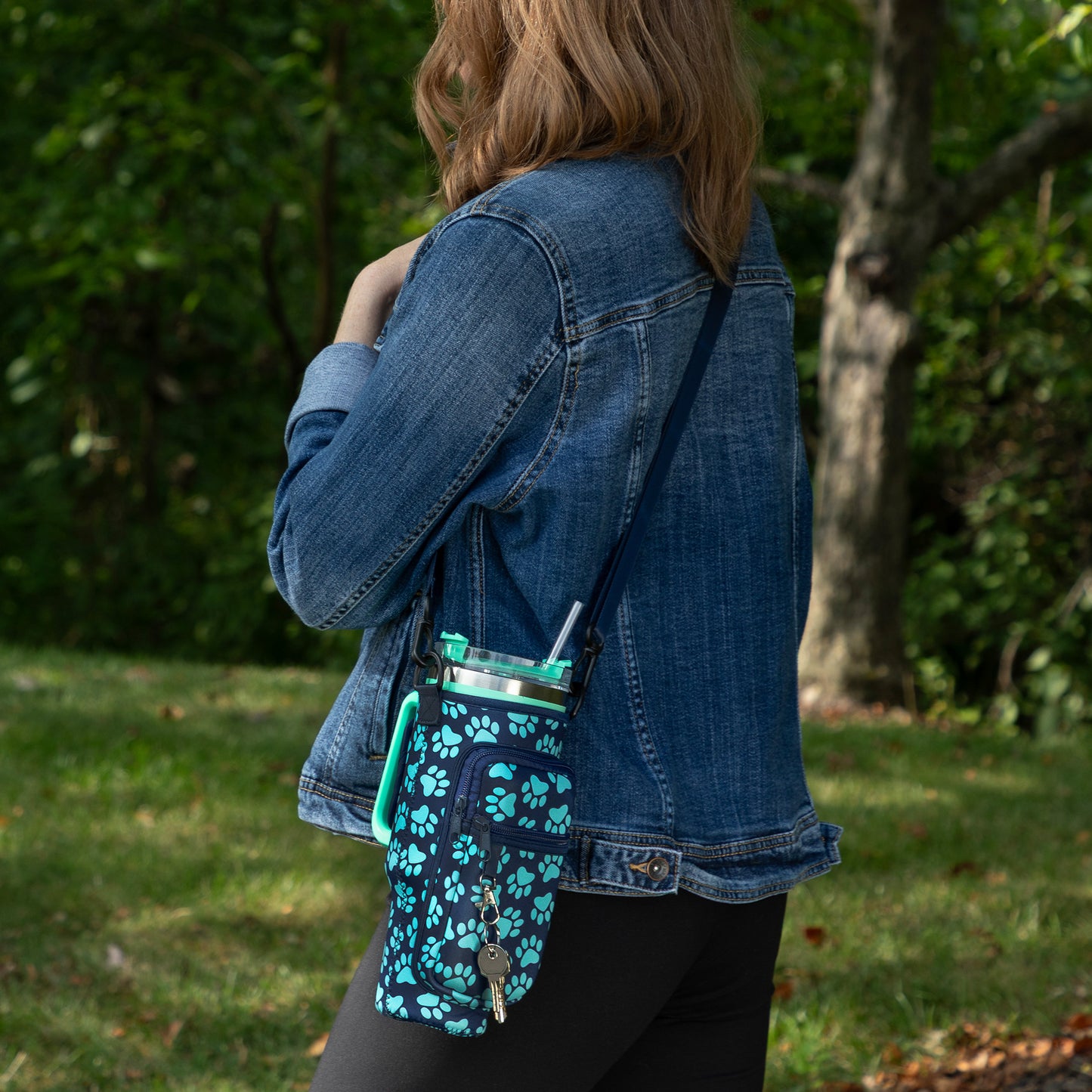A woman wearing a denim jacket stands outdoors in a grassy area with trees in the background, carrying a small crossbody tumbler bottle. The bottle has a dark navy base with a bright turquoise paw-print pattern and a matching turquoise loop handle.