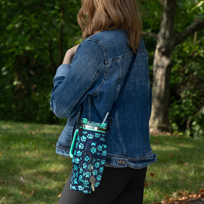 A woman wearing a denim jacket stands outdoors in a grassy area with trees in the background, carrying a small crossbody tumbler bottle. The bottle has a dark navy base with a bright turquoise paw-print pattern and a matching turquoise loop handle.