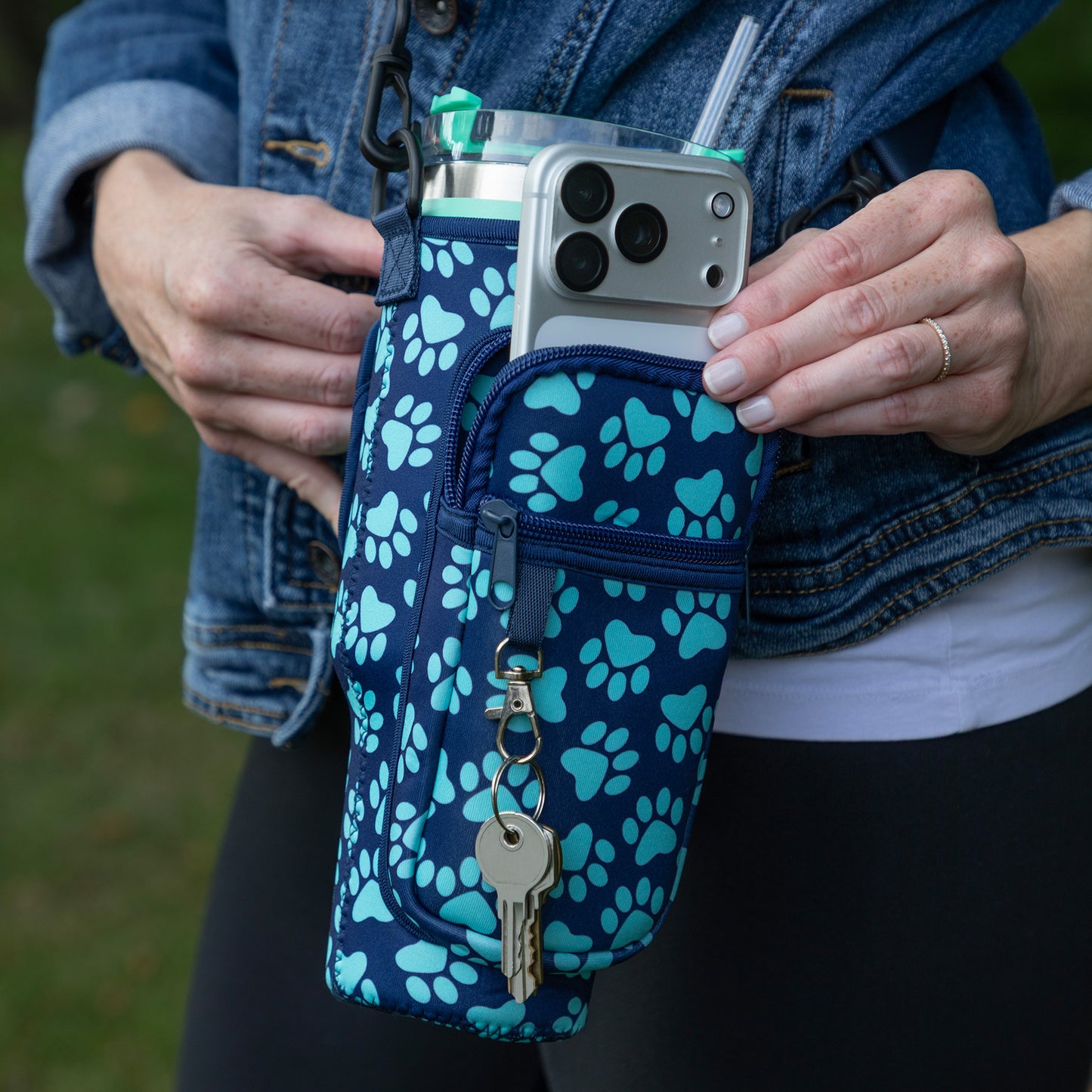 A close-up of a person wearing a blue and teal paw-print patterned phone pouch, holding a silver smartphone in it. The pouch has a front zip pocket with a keychain and a side loop holding a clear water bottle with a green cap.