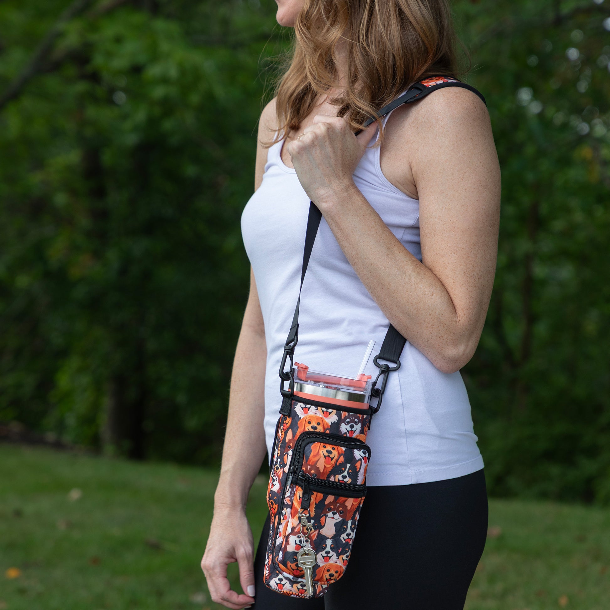 A woman wearing a white tank top and black leggings is shown from the side outdoors, wearing a small crossbody pouch bag with a black and orange dog pattern and a clear water bottle inside.