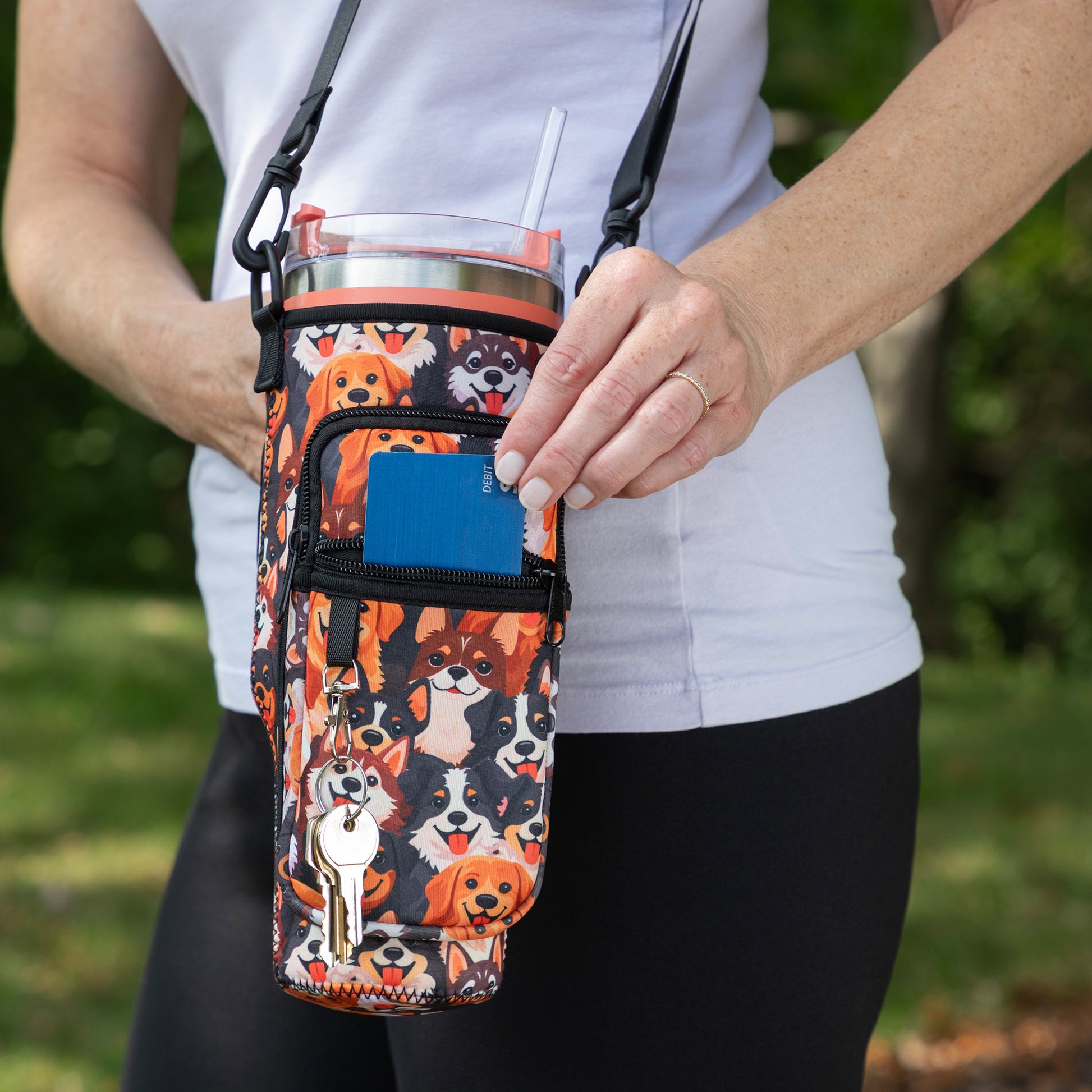 A person outdoors holding a dog-print insulated tumbler bag with a shoulder strap and front zip pocket. The bag features multiple dog illustrations in orange, black, white, and tan, with a small dog-shaped keychain attached.