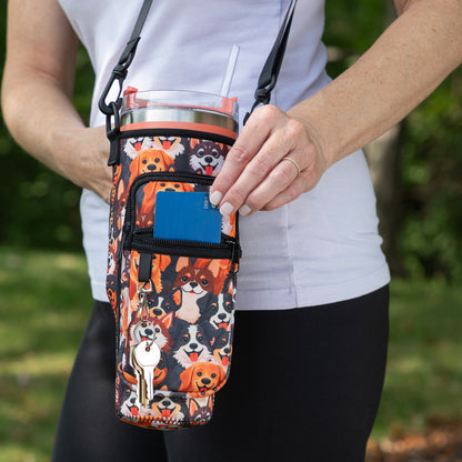 A person outdoors holding a dog-print insulated tumbler bag with a shoulder strap and front zip pocket. The bag features multiple dog illustrations in orange, black, white, and tan, with a small dog-shaped keychain attached.
