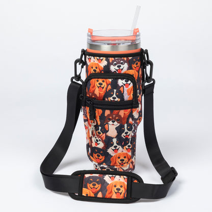 A black insulated tumbler sleeve with an all-over dog print, shown with a clear-lidded tumbler and straw. The sleeve includes a front zip pocket and an adjustable shoulder strap, photographed on a clean white studio background.