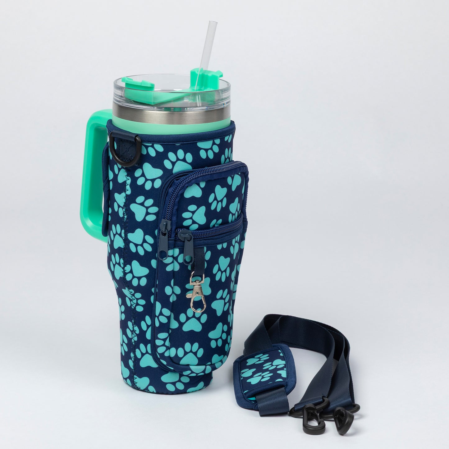 A navy blue insulated tumbler with a mint green handle and lid, covered by a matching navy sleeve printed with mint green paw prints. The tumbler has a clear lid with a mint green straw, and a separate mint green-and-navy shoulder strap is placed beside it on a light gray studio background.