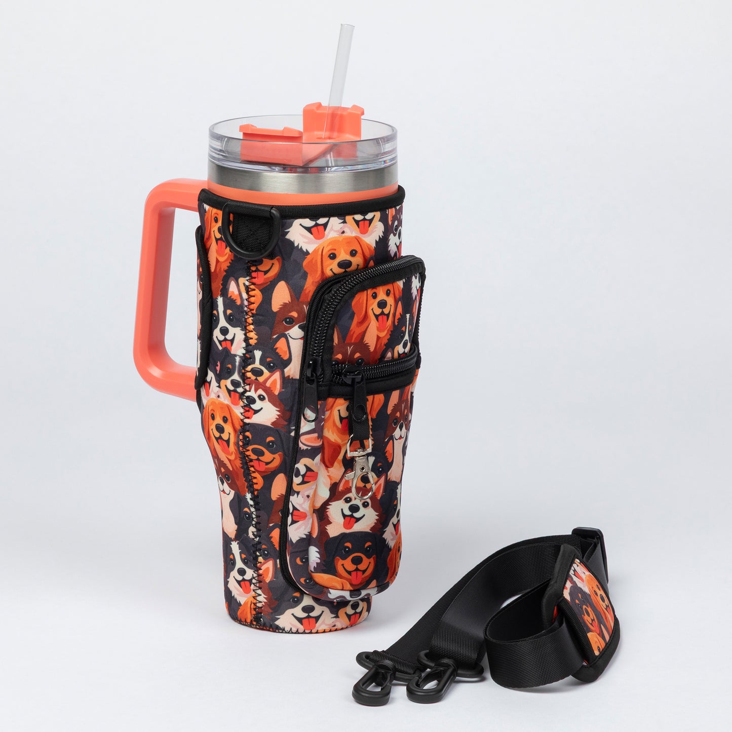 A stainless steel handled tumbler with a clear lid and orange straw, wrapped in a black sleeve printed with a collage of dog faces in orange, cream, and black. A matching black shoulder strap with an orange-and-black printed patch lies on the white studio background.