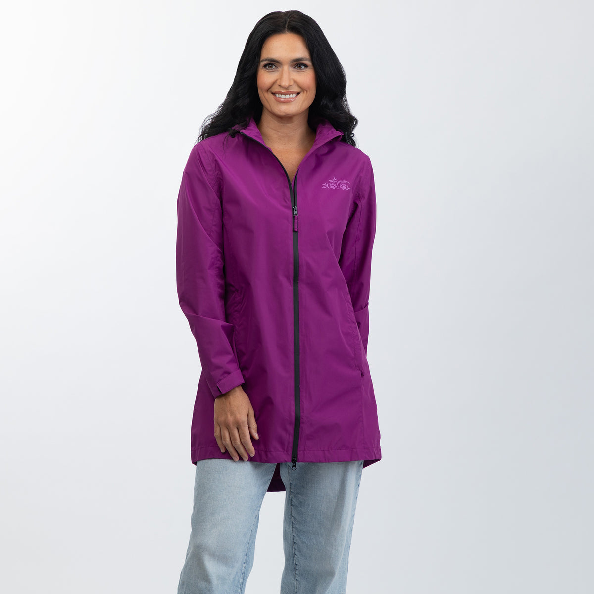 A woman wearing a purple full-zip jacket with a small white embroidered logo on the left chest, paired with light blue jeans, standing against a light gray studio background.
