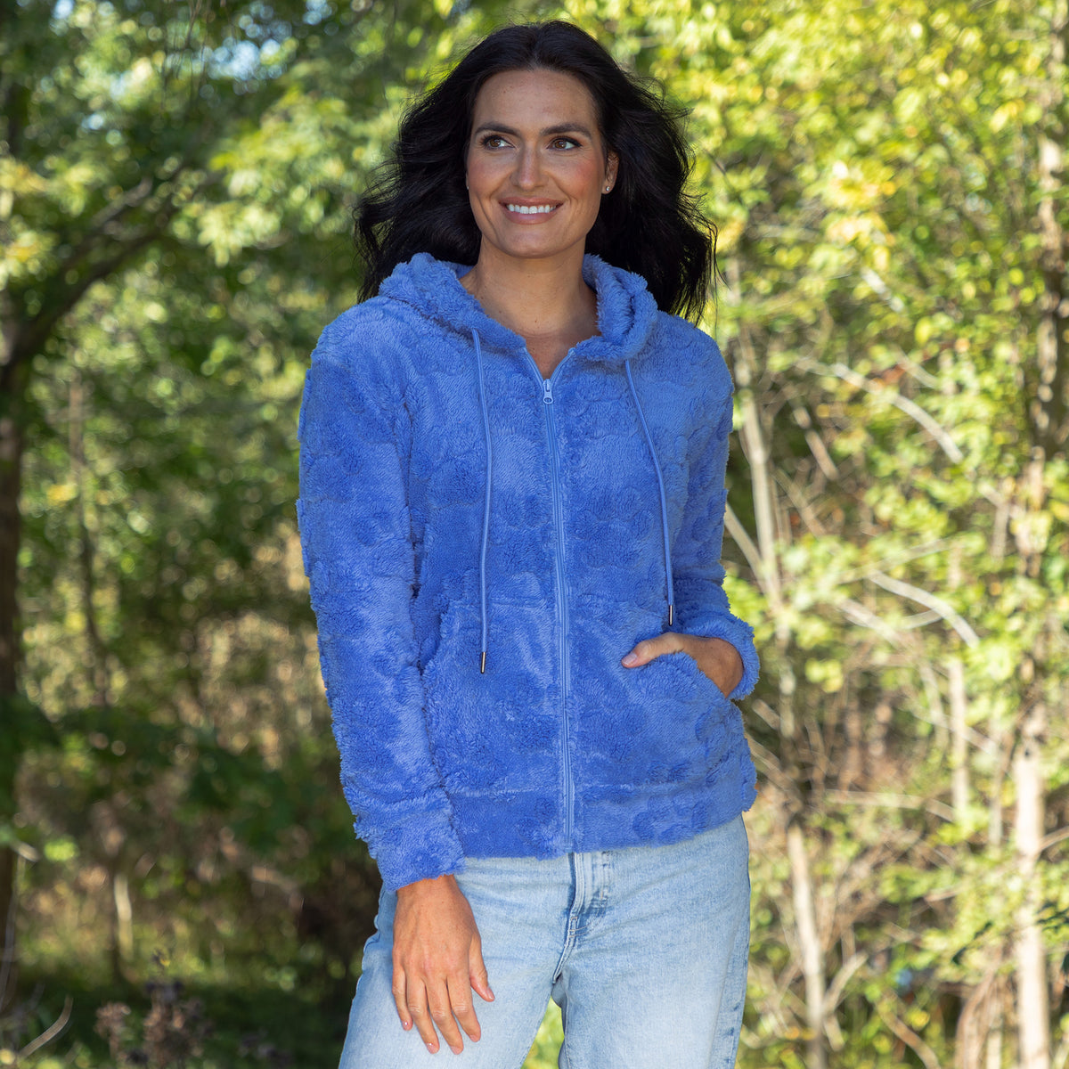 A woman wearing a fuzzy blue zip-up hoodie and light-wash jeans, standing outdoors in front of a blurred green forest background.