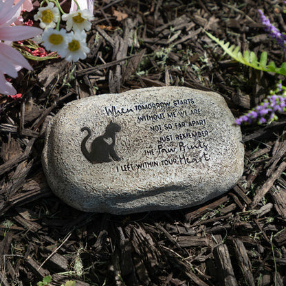 Decorative stone with a cat silhouette and engraved text on a mulched garden bed with flowers.
