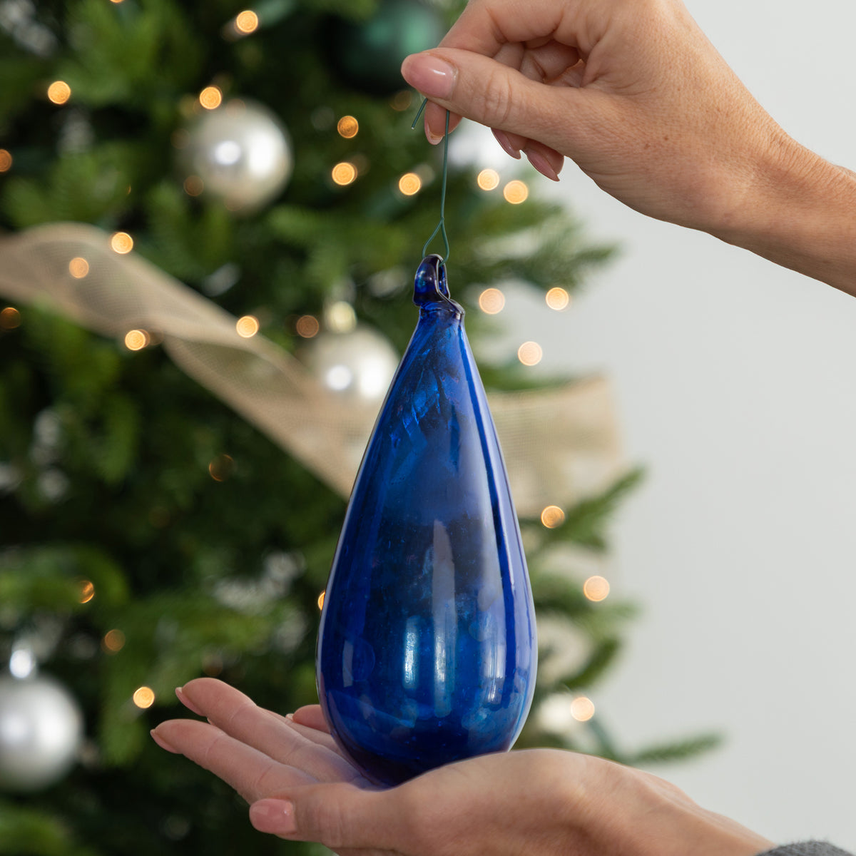 A person holds a large translucent cobalt blue glass teardrop Christmas ornament in front of a softly blurred decorated Christmas tree with lights and ornaments.