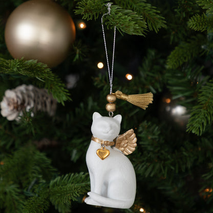 A white cat Christmas ornament with gold accents, including a collar, heart charm, wings, and tassel, hanging on a decorated Christmas tree.