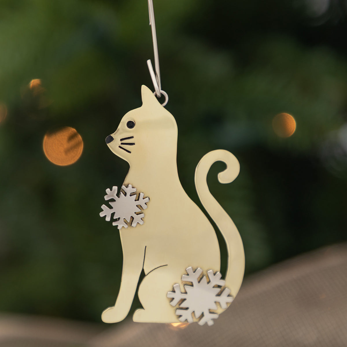 A light cream-colored metal cat Christmas ornament with silver snowflake accents, hanging against a dark green blurred background with warm bokeh lights.