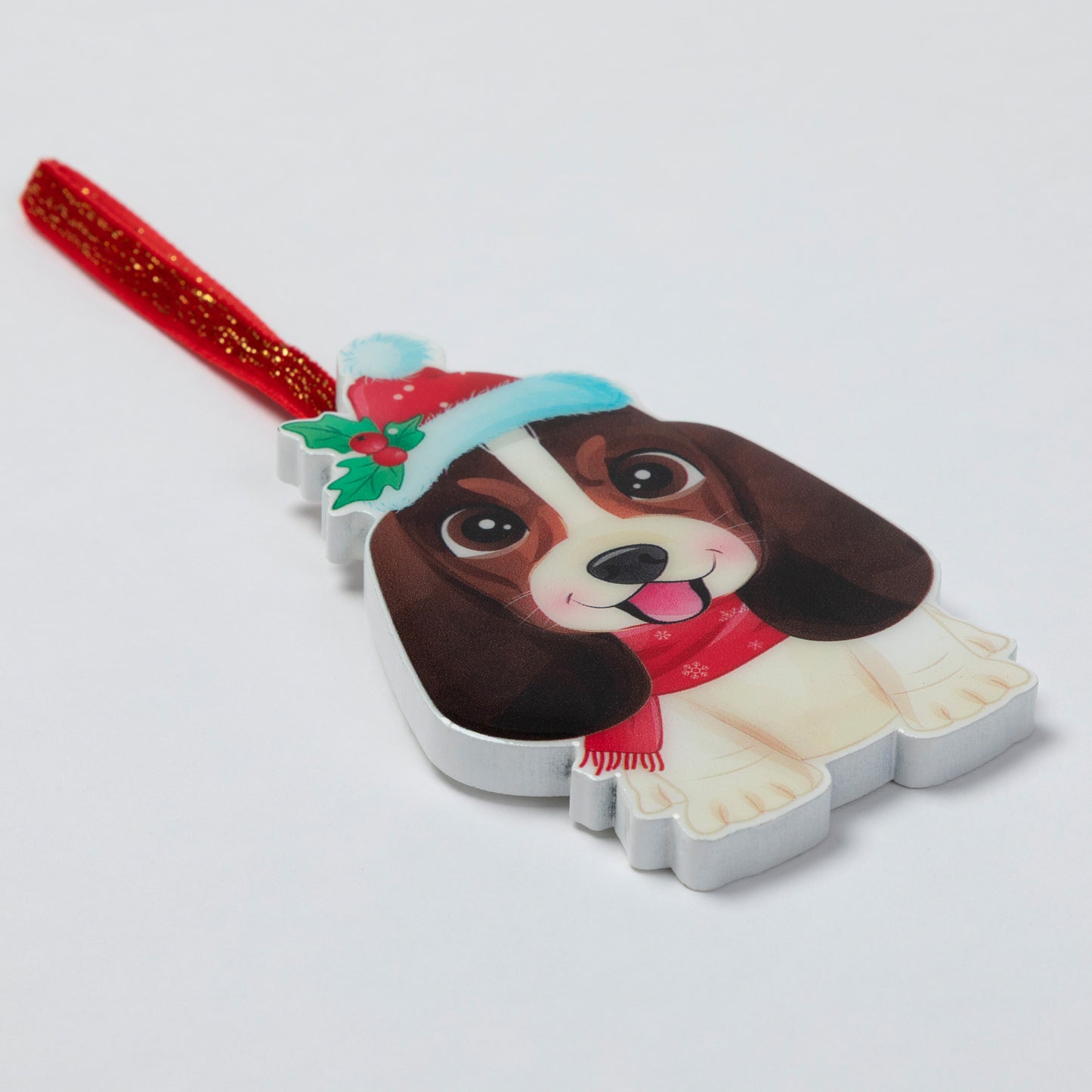 A Christmas dog-shaped ornament featuring a brown and cream dog wearing a red Santa hat and scarf, with a red glittery hanging loop, photographed on a light gray background.