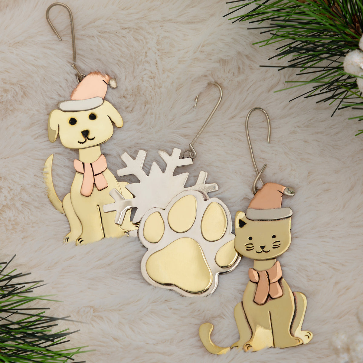 Three Christmas ornaments shaped like a dog, a cat, and a paw print, decorated with pastel colors and hanging hooks, displayed on a white faux fur background with pine branches in the corners.