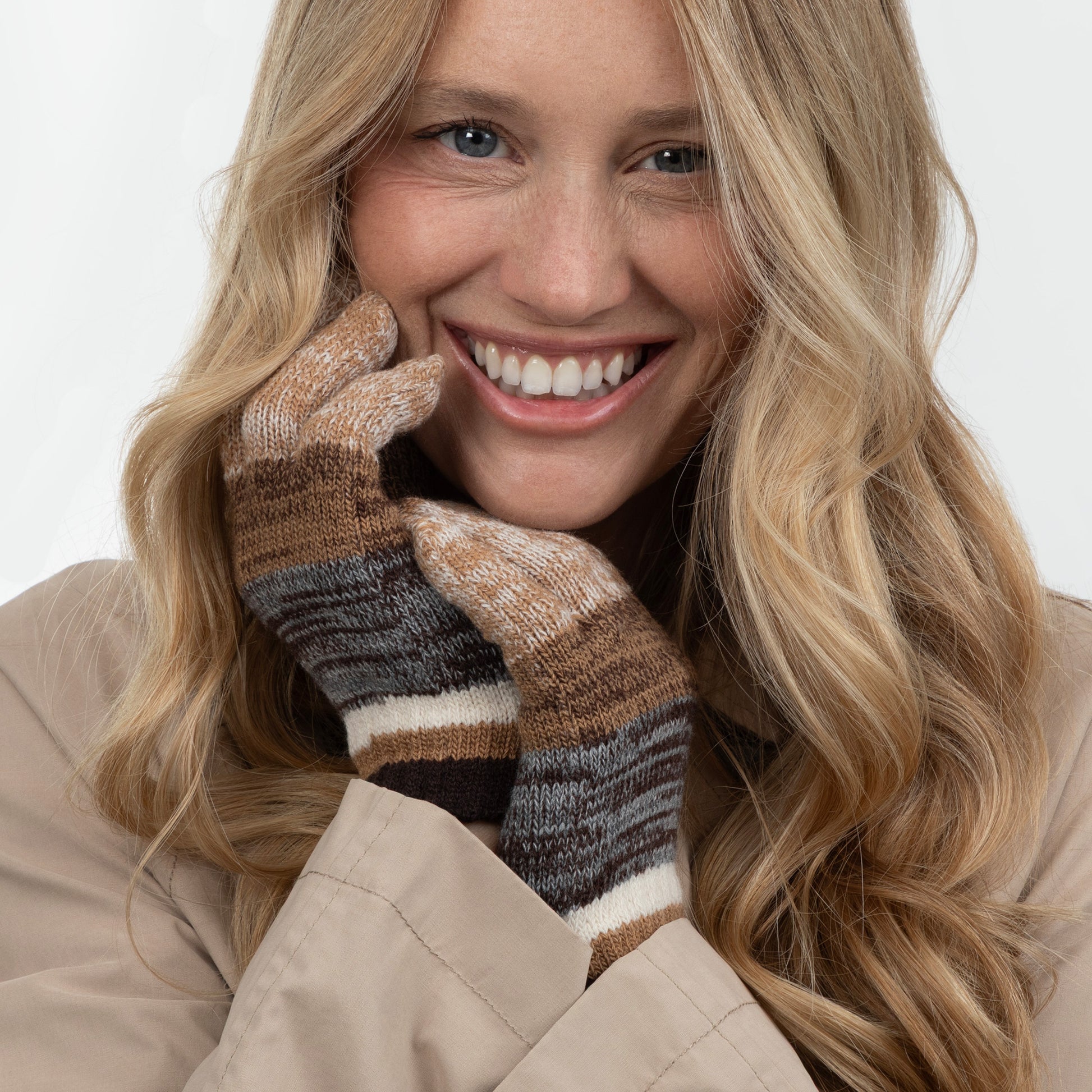 A smiling blonde woman wearing a pair of knit striped gloves in brown, beige, gray, and white, paired with a beige coat against a plain light background.