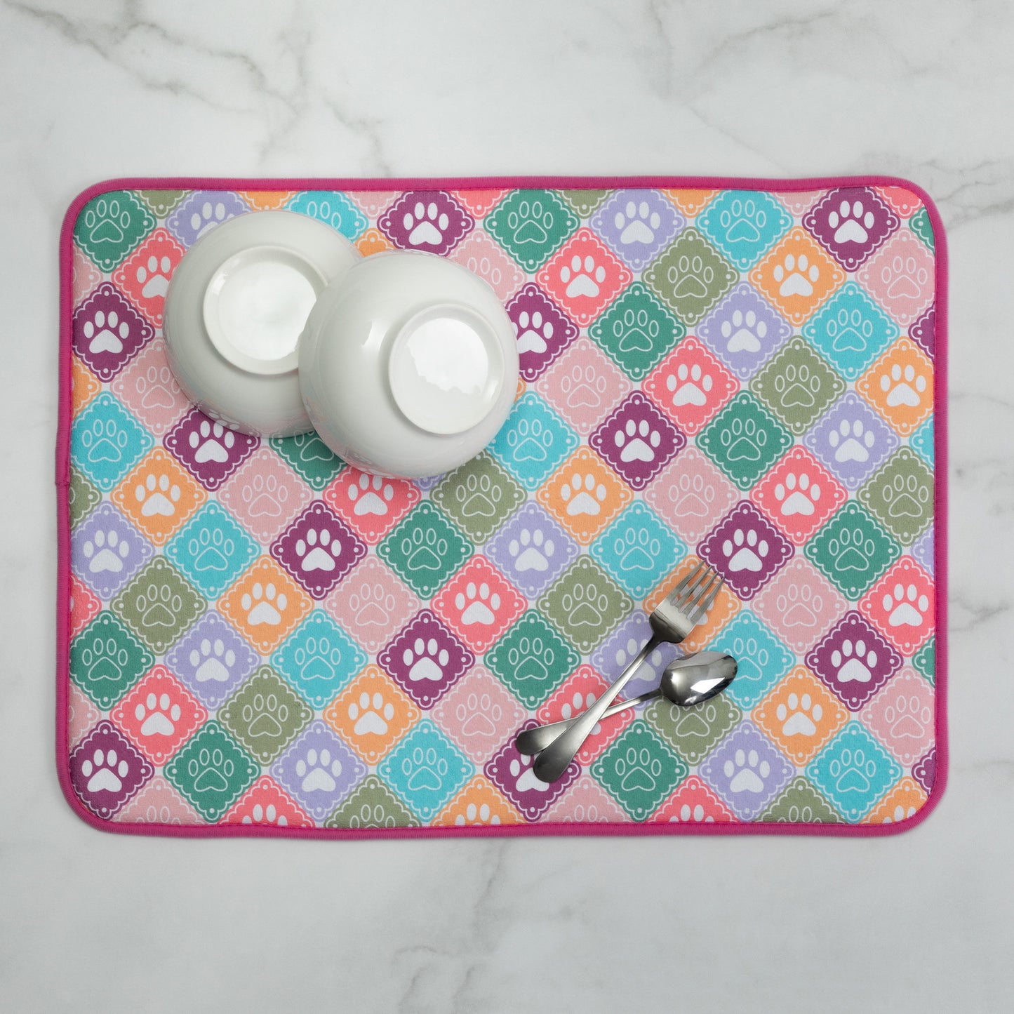 A rectangular dish drying mat with a colorful diamond pattern featuring paw prints, styled with two white bowls and metal utensils on a white marble surface.