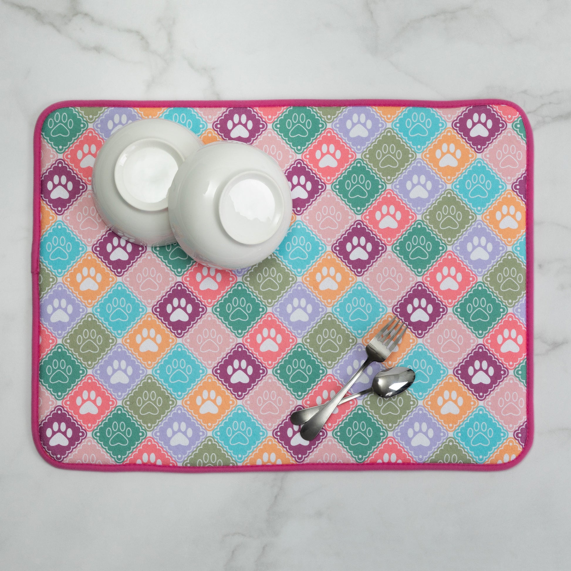 A rectangular dish drying mat with a colorful diamond pattern featuring paw prints, styled with two white bowls and metal utensils on a white marble surface.