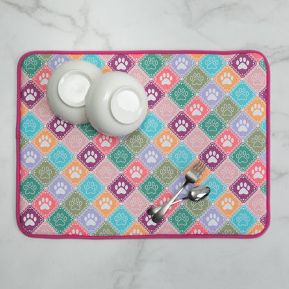 A rectangular dish drying mat with a colorful diamond pattern featuring paw prints, styled with two white bowls and metal utensils on a white marble surface.
