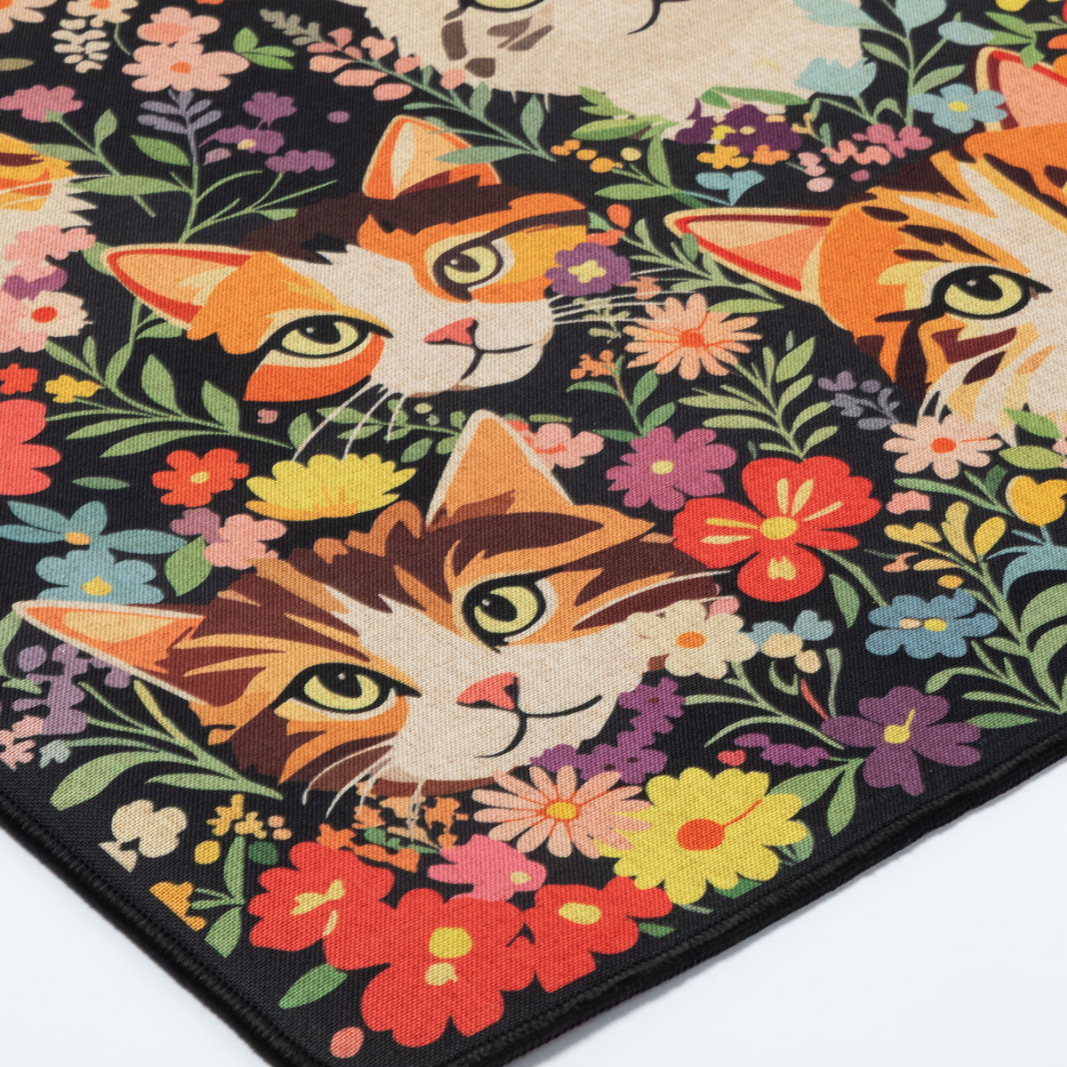 A close-up of a colorful area rug featuring three orange tabby cat faces surrounded by a dense pattern of multicolored flowers on a black background.