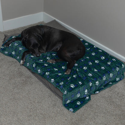 A black dog rests on a rectangular dog bed with a green plaid blanket featuring white paw prints, placed on a carpeted floor in a room corner.