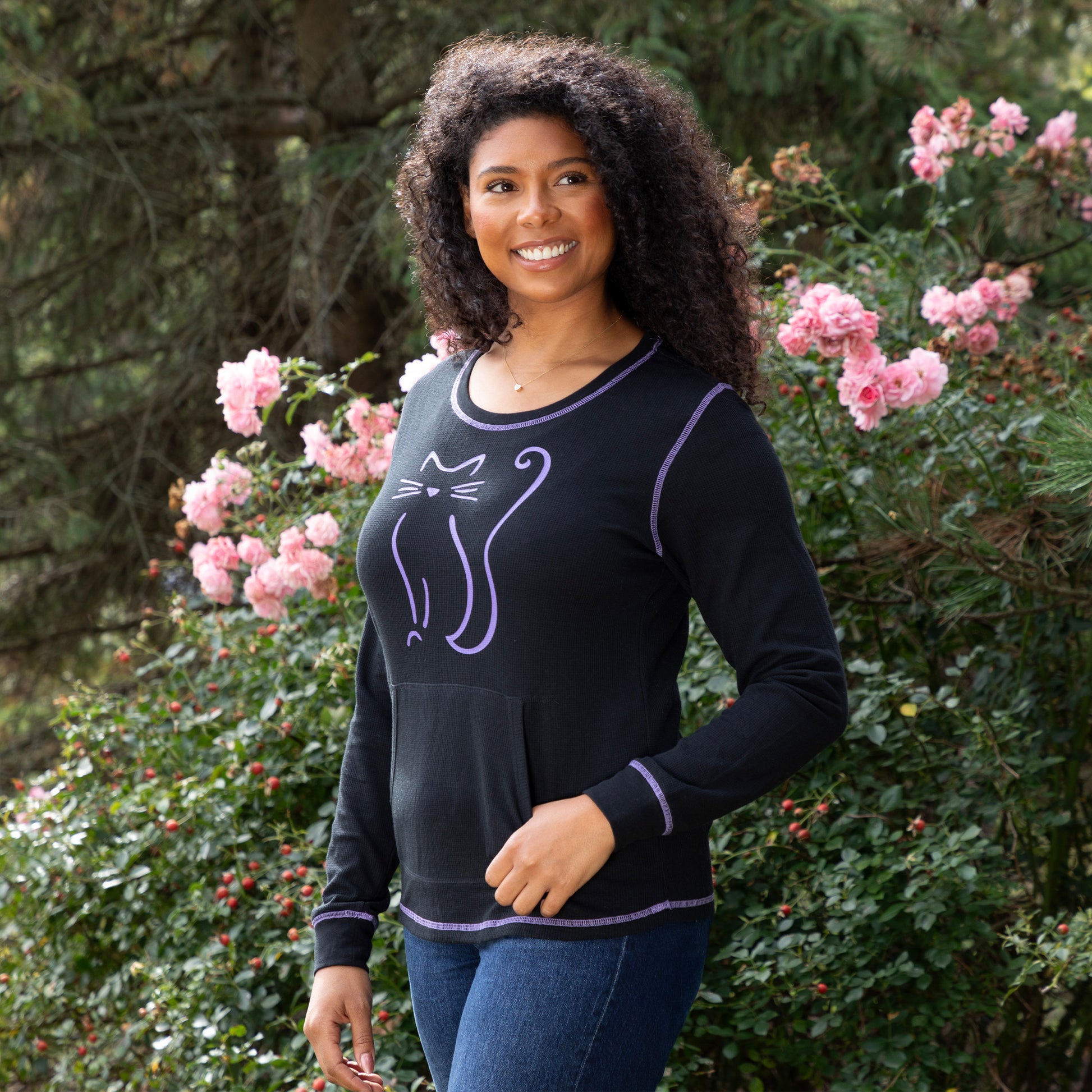 Smiling woman outdoors in a cat-themed black thermal long sleeve top surrounded by colorful flowers.
