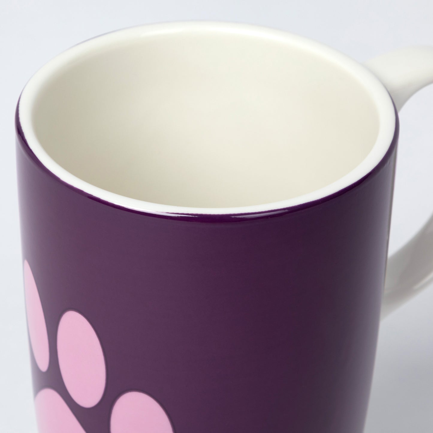 A close-up of a ceramic mug with a deep purple exterior, decorated with a pink paw print pattern, and a white interior and handle.