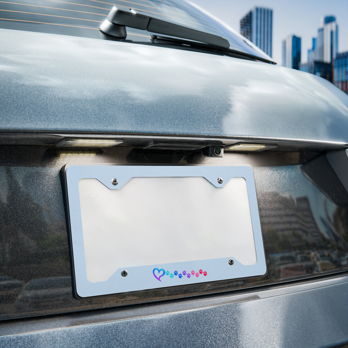 A silver license plate frame with a colorful heart pattern along the bottom, mounted on the rear bumper of a gray car with a city skyline in the background.