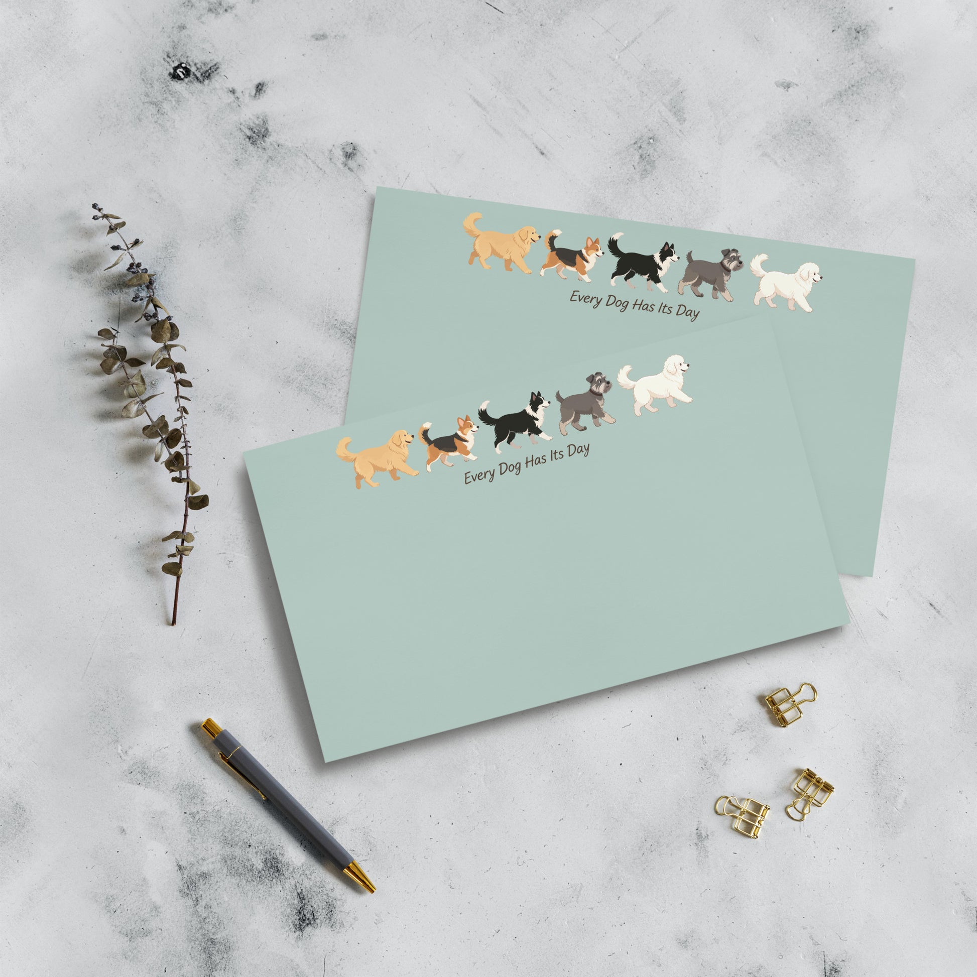 Two 10x6 mint green notepads with illustrations of various dog breeds and the text 'Every Dog Has Its Day' are arranged on a light gray marble surface with a pen, paperclip clips, and a sprig of eucalyptus.