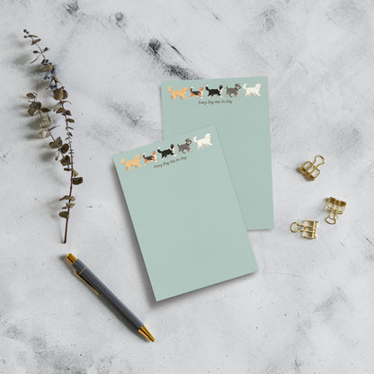 Two 4x6 pastel green notepads with a row of dog illustrations and the text 'Every dog has its day' on the top page, arranged on a light marble surface with a pen, dried stems, and gold binder clips.