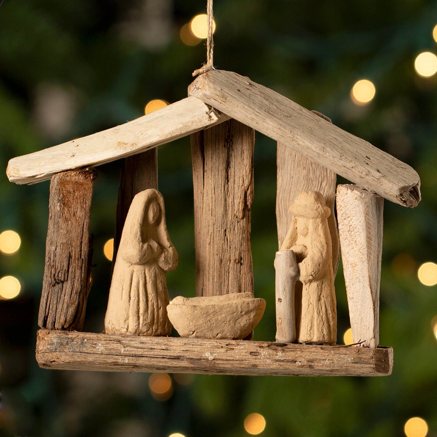 A handmade artisan recycled driftwood Christmas ornament featuring a nativity scene, manger, hanging against a blurred background of Christmas lights.